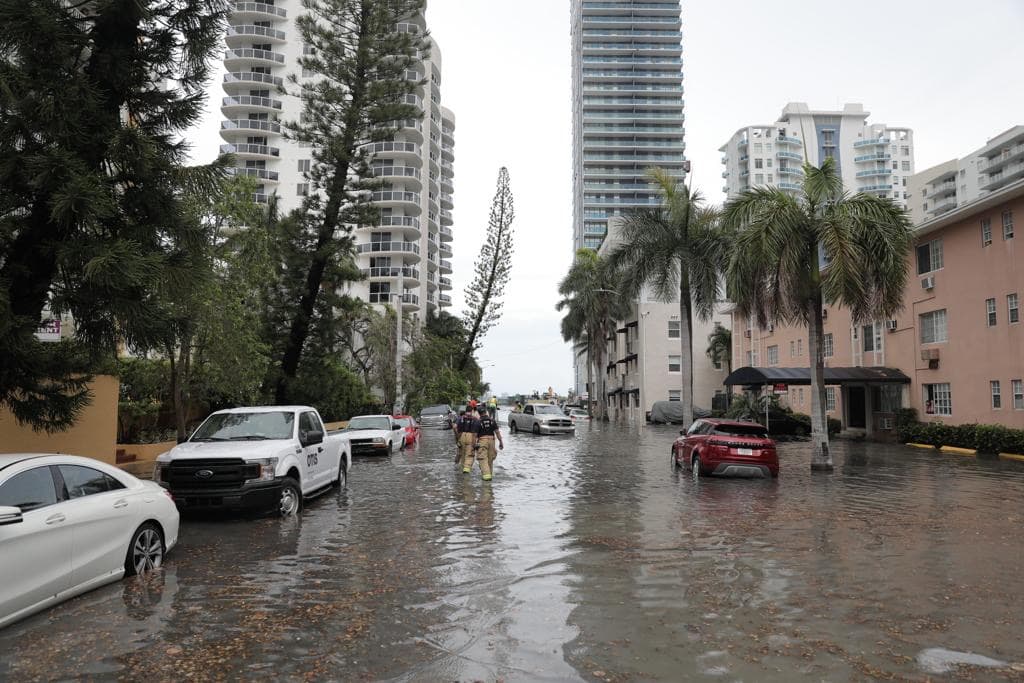 A flooded South Florida street during king tide season, with cars parked in standing water between apartment buildings and palm trees.